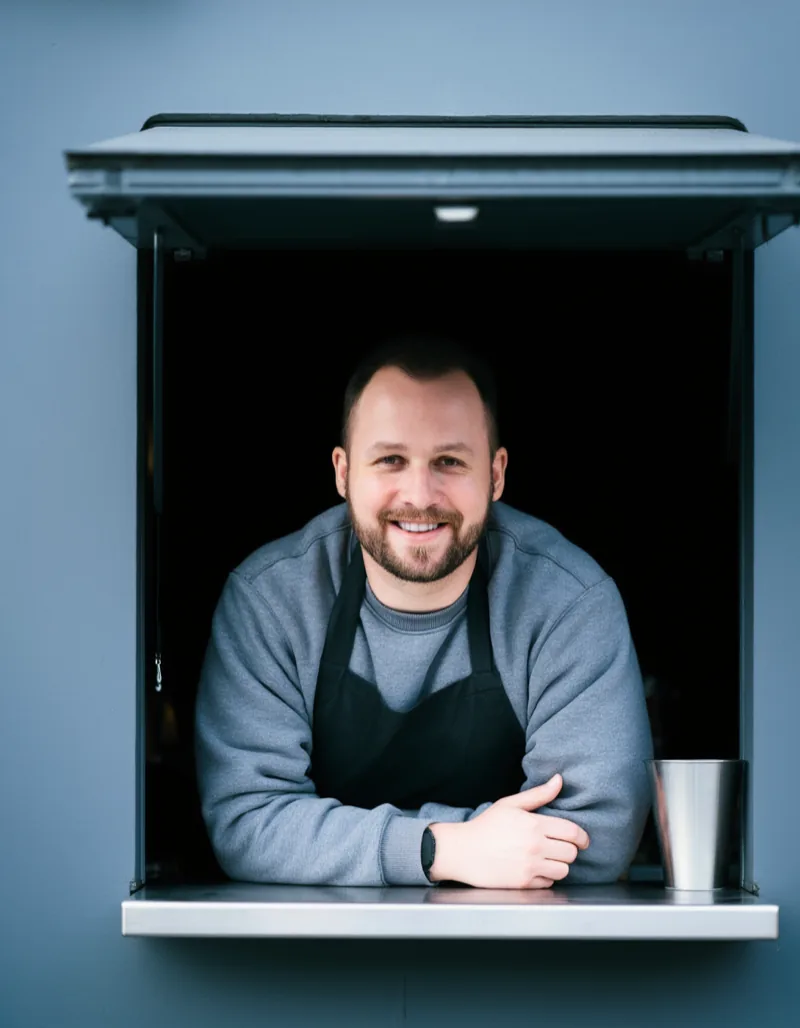 A barista in a dark apron leaning out through the square serving hatch, arms resting on the steel ledge