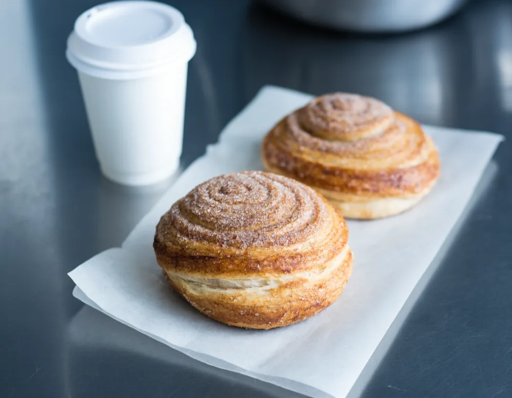 Two cinnamon pastries on a sheet of greaseproof paper on the steel counter, next to a lidded paper coffee cup