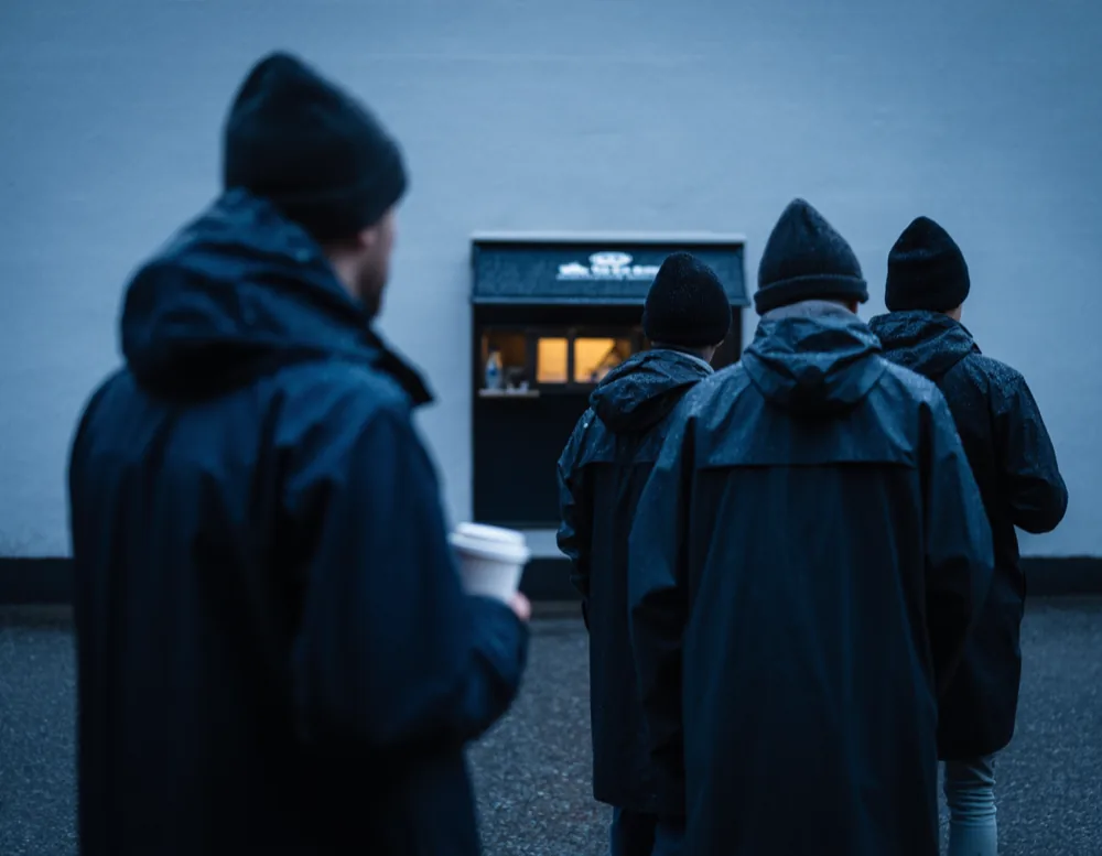 A short queue of commuters in dark rain jackets waiting on the wet pavement at the hatch